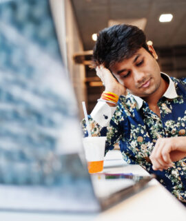 Young male indian freelancer sitting open laptop computer in fast food cafe, handsome asian man looking at watches during work on net-book in comfortable coffee shop.